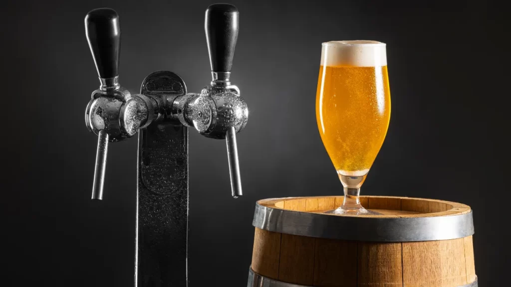 Imagem mostrando uma torneira de chope metálica com duas saídas, ao lado de um barril de madeira com um copo cheio de chope dourado com espuma, sob um fundo escuro e elegante.