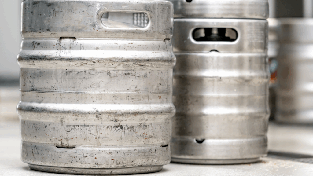 Barris de cerveja em inox usados, com marcas de desgaste, posicionados em um ambiente industrial.